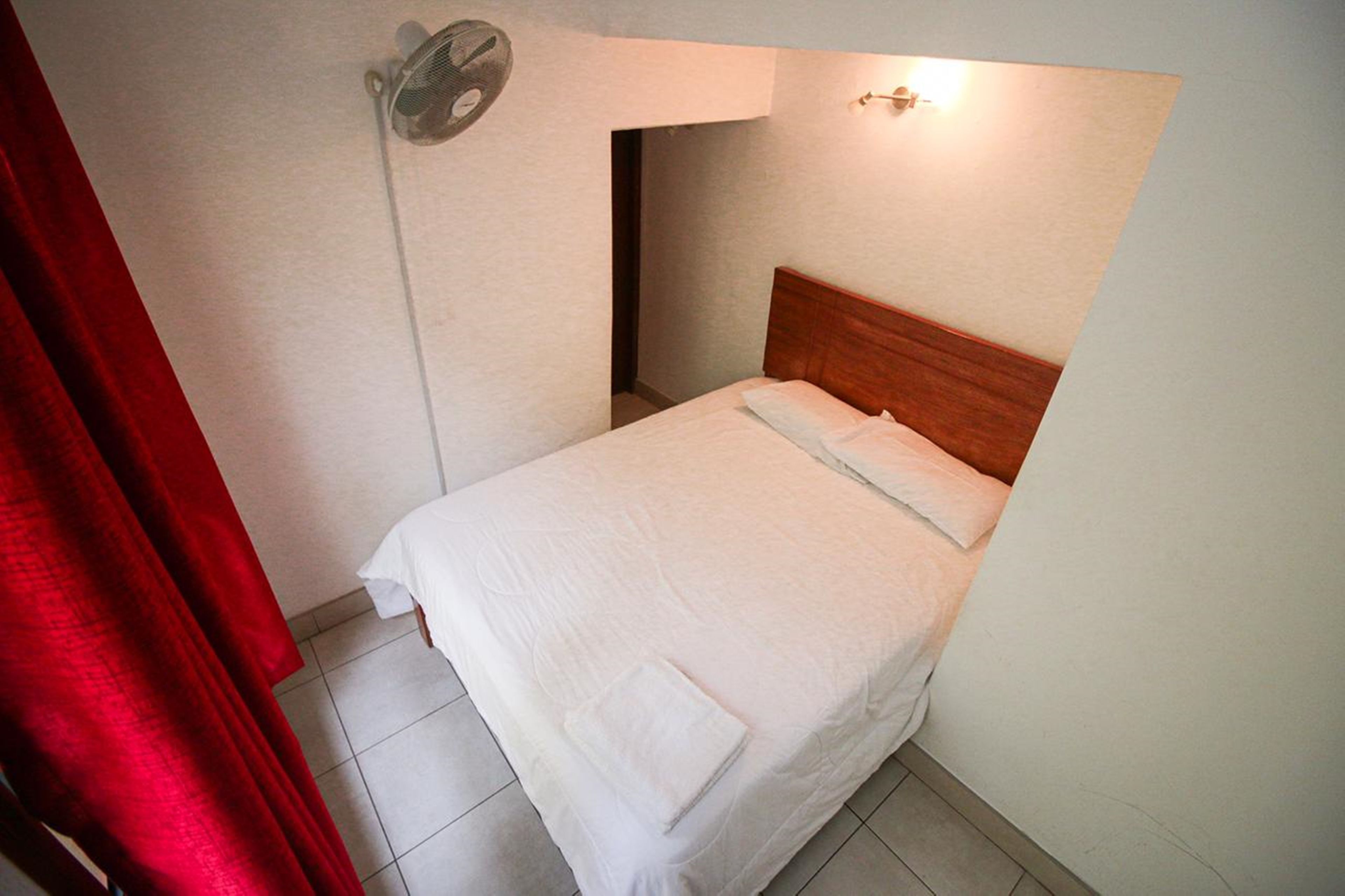 Un lit bien fait, avec une tête de lit en bois, dans une petite pièce bien éclairée et dotée d’un sol carrelé.