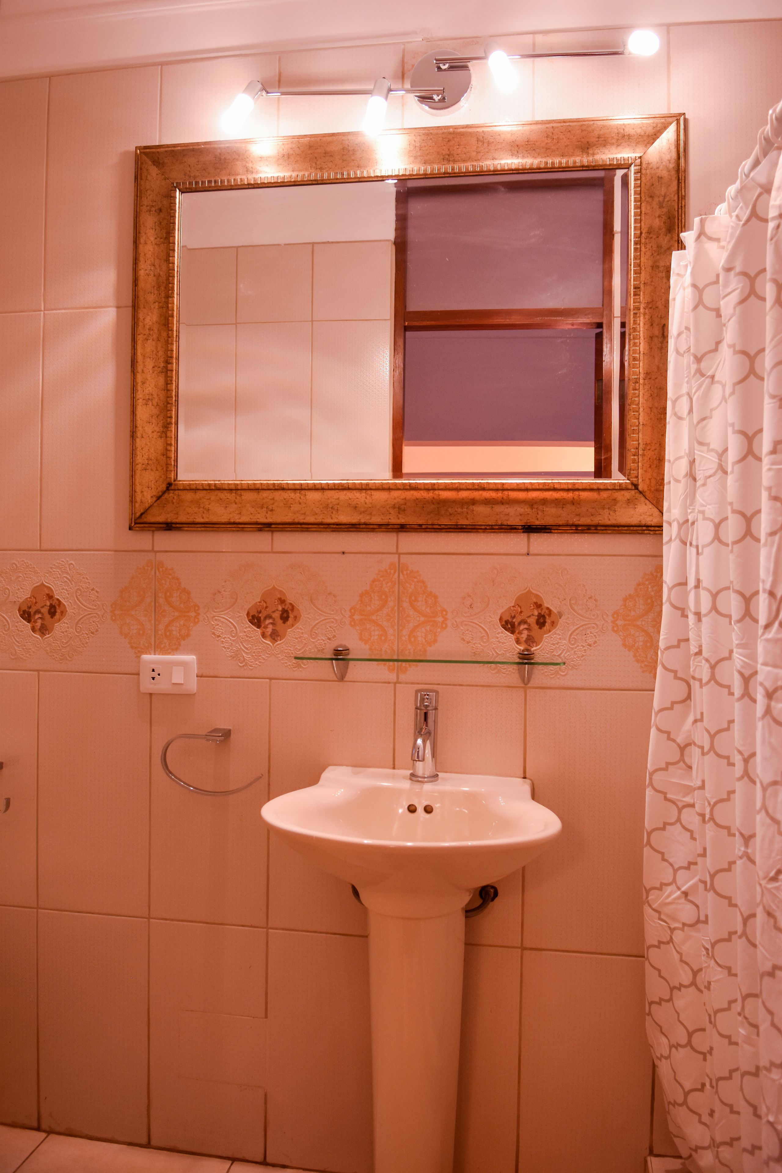 A bathroom with a pedestal sink, a mirror with lights, patterned tiles, and a shower curtain.