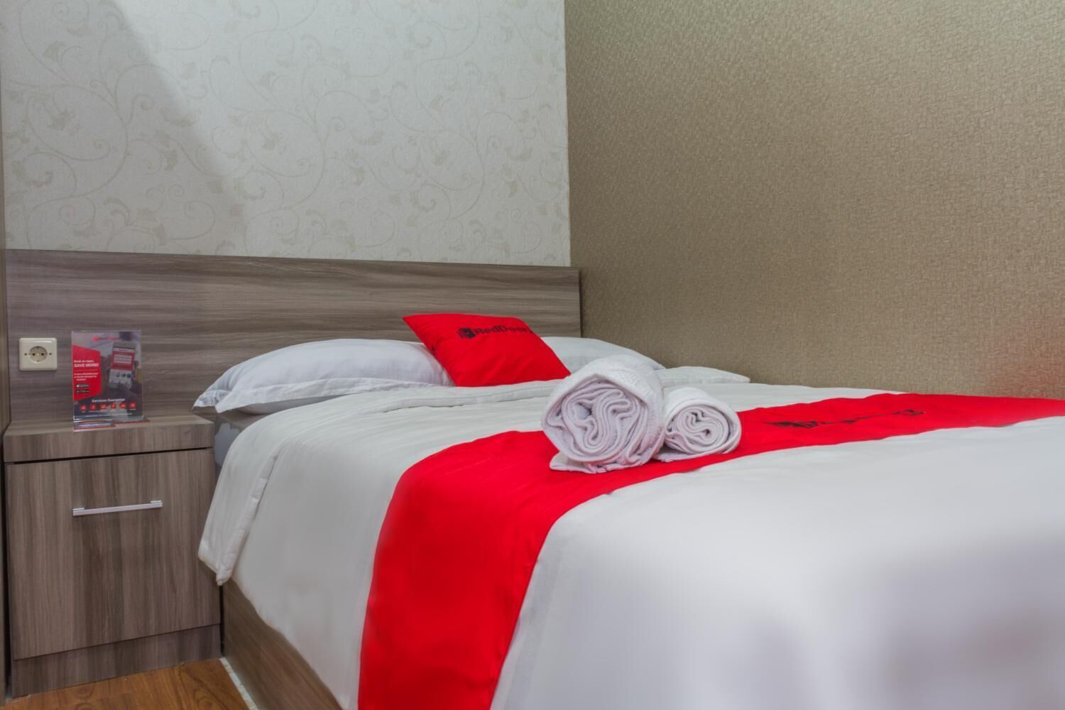 A neatly made bed with white and red bedding, a red pillow, and rolled towels. There is a small table with a red object on it, and a wall with a subtle pattern.