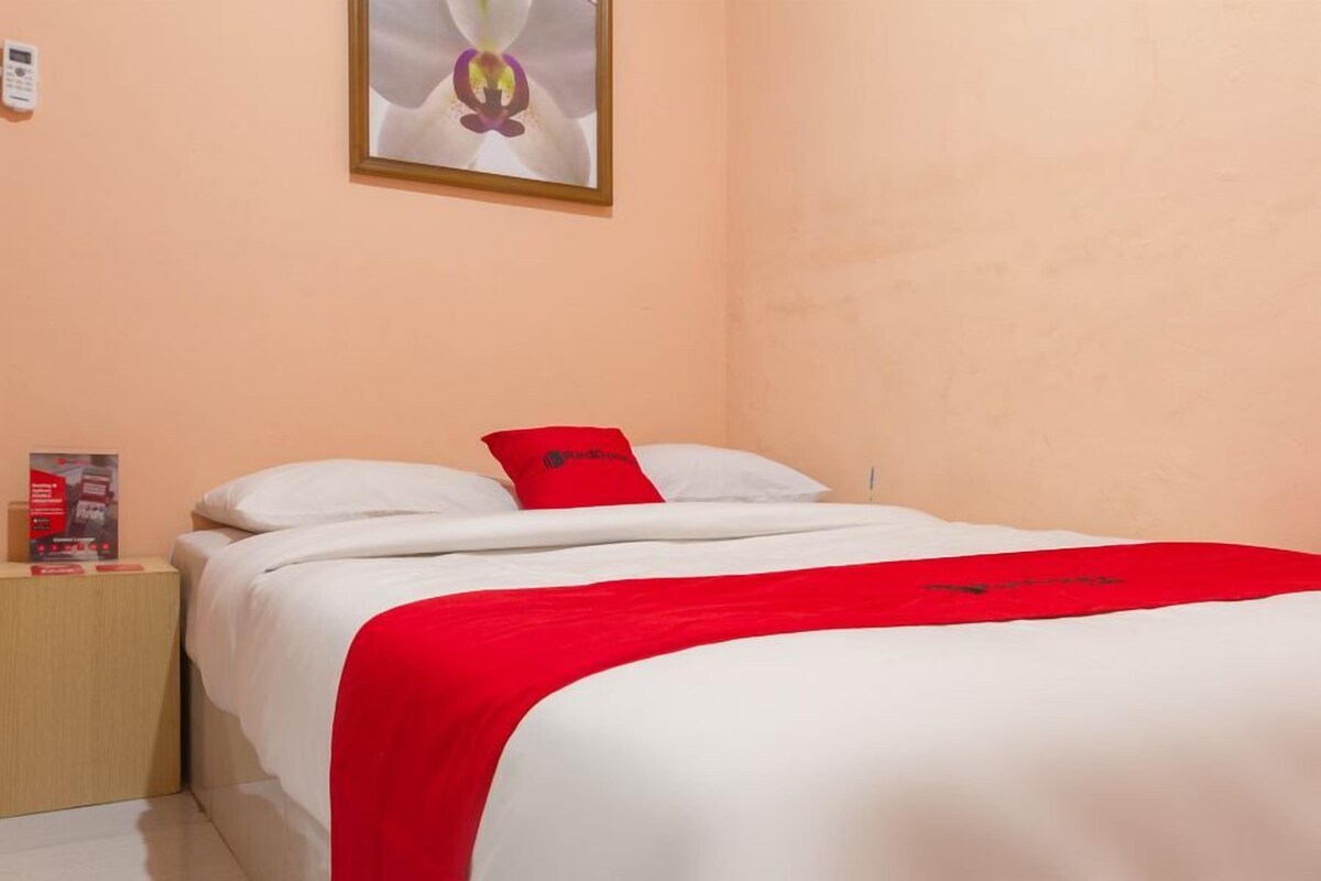 Un lit bien fait, avec une literie blanche et rouge, une petite table de chevet sur laquelle se trouve un objet rouge, et un tableau encadré accroché au mur.