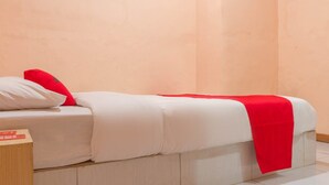 Un lit simple avec une literie blanche et une couverture rouge, un oreiller blanc et un oreiller rouge, le tout adossé à une tête de lit en bois, dans une pièce aux murs de couleur pêche.