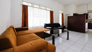 Una sala de estar con un sofá marrón, un sofá de cuero negro y una mesa de centro de vidrio. Hay dos ventanas con cortinas marrones, un armario de madera oscura y una zona de cocina al fondo.