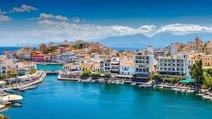 Un poble costaner amb edificis al front marítim, una marina amb vaixells i muntanyes al fons.