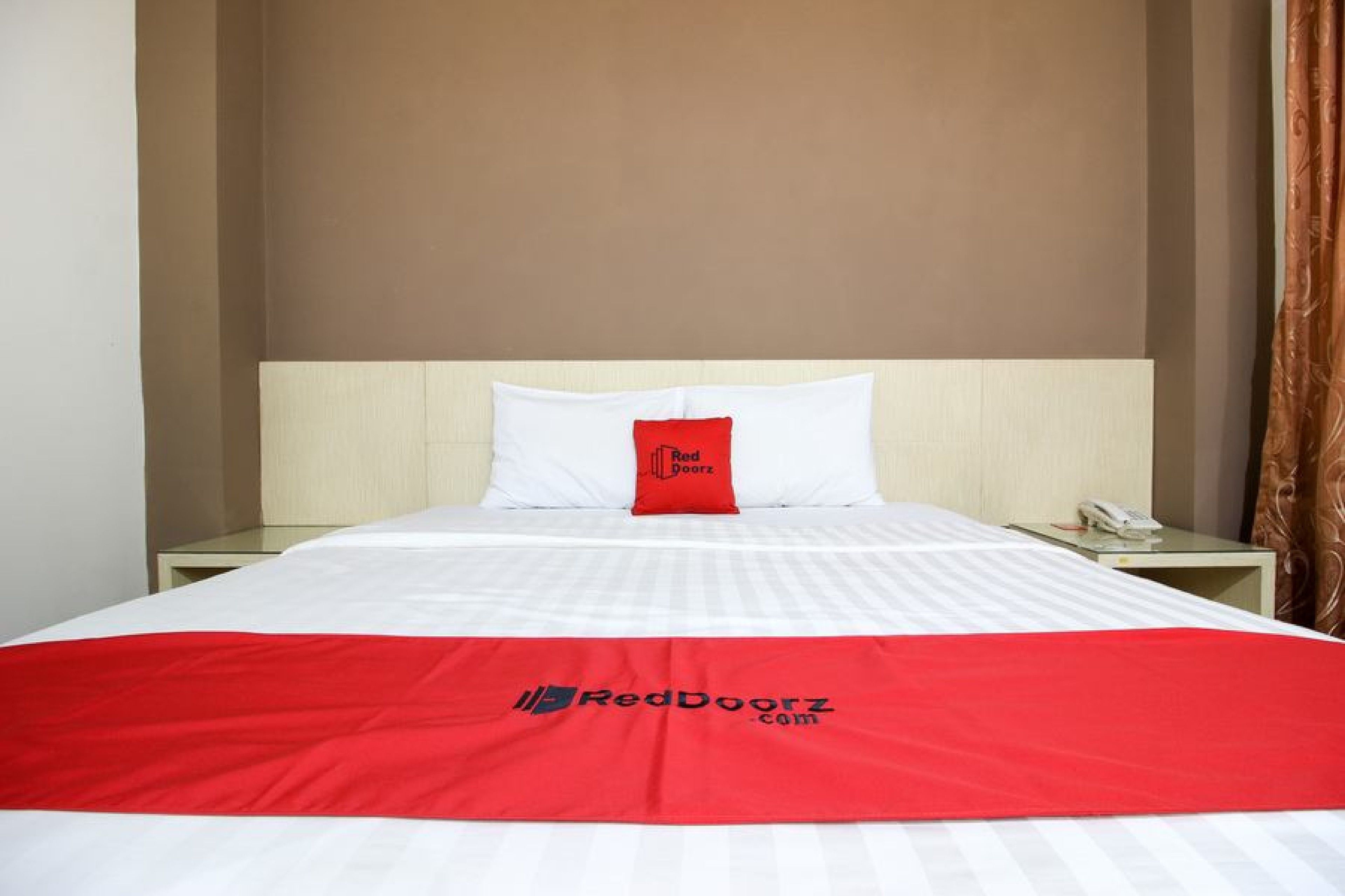 A hotel room with a neatly made bed, a red and white bedspread, a pillow with a red logo, and a bedside table with a phone.