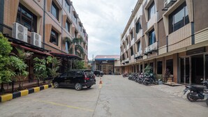 Un espace de stationnement où sont garées de nombreuses voitures et motos, entouré d’immeubles dotés de climatiseurs et agrémenté de plantes en pot.