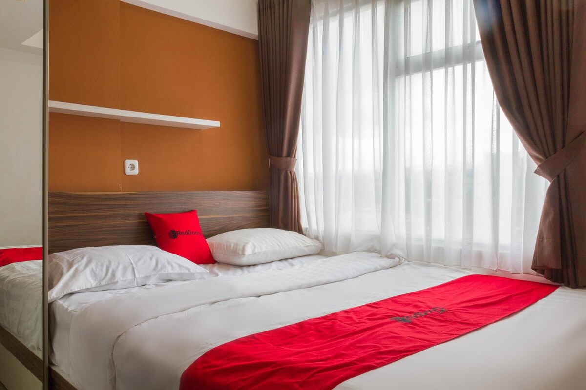 Une chambre d’hôtel avec un lit, un jeté rouge, du linge de lit blanc, une tête de lit et des rideaux transparents.