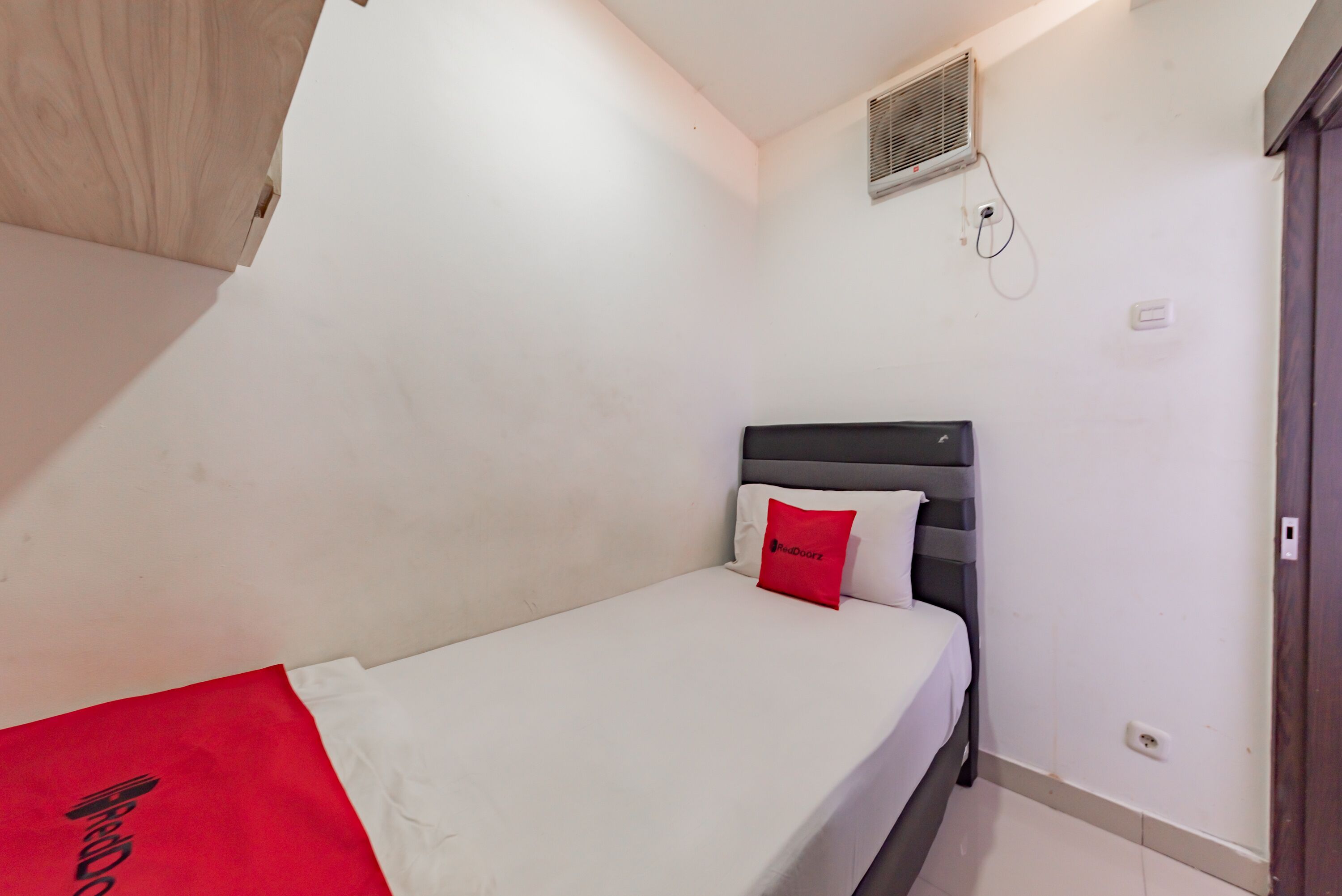 Una cama individual con cabecera gris, colchón blanco y almohada roja. Se aprecia una pared blanca con un respiradero y un enchufe.