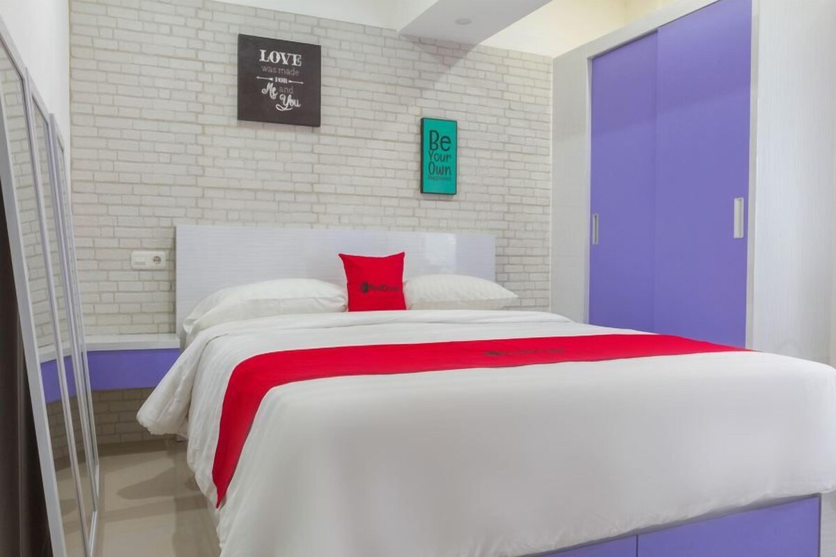 Une chambre avec un mur en briques blanches, un lit avec une literie blanche et un oreiller rouge, une porte bleue et deux œuvres d’art murales inspirantes.