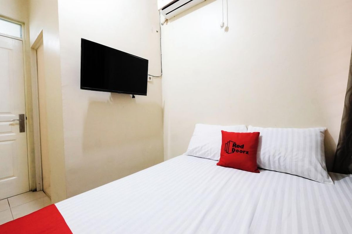 A hotel room with a bed, a flat-screen TV mounted on the wall, and a red pillow with text.