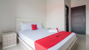 A neatly made bed with white bedding and a red blanket, a wooden nightstand with drawers, and a dark wooden door. - RedDoorz Syariah near Tugu Juang Jambi (Jambi)
