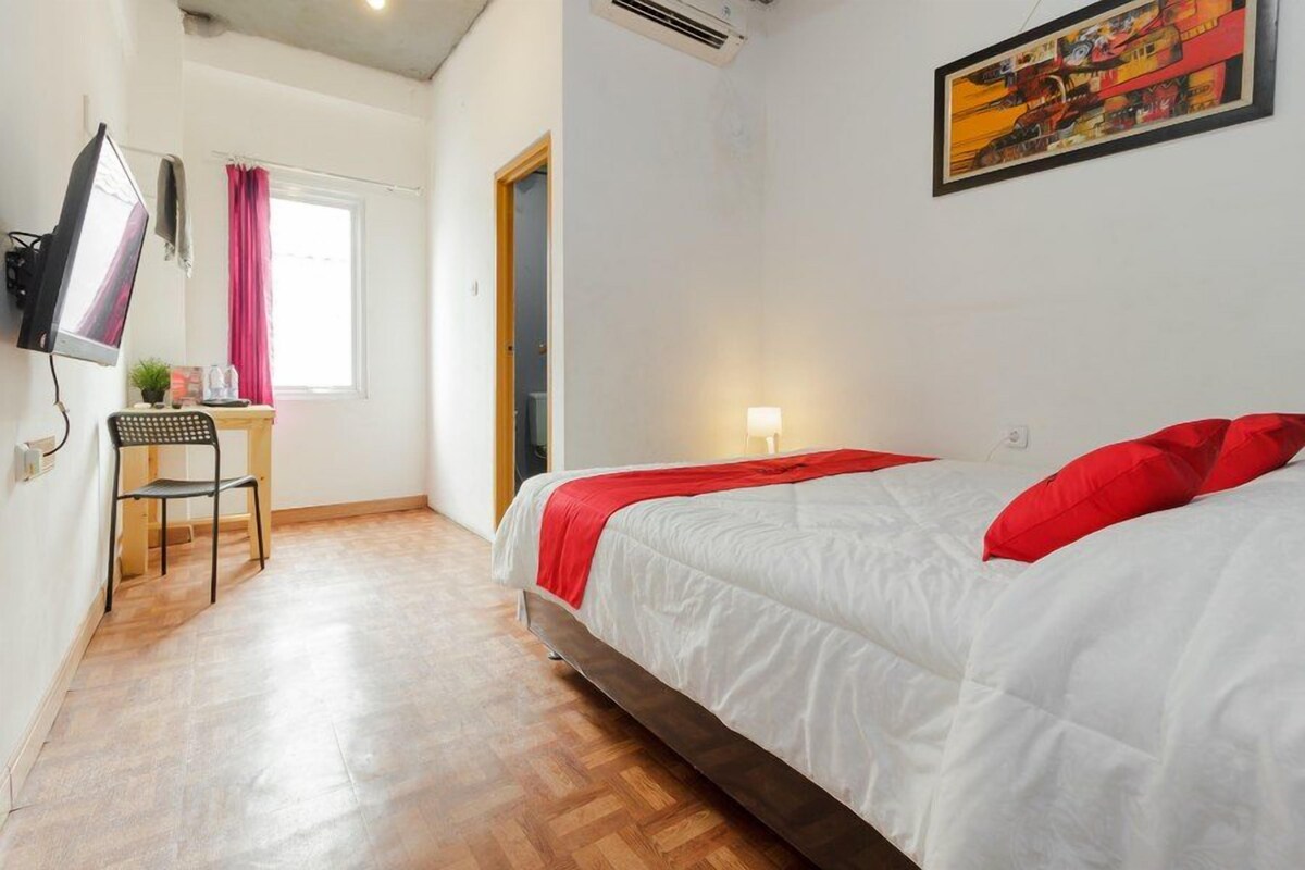 A bedroom with a bed, a desk, a chair, a TV, and a painting on the wall.