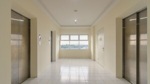 A hallway with two elevators, a large window with a view, and a tiled floor. - RedDoorz Apartment @ Dramaga Tower (Ciampia)