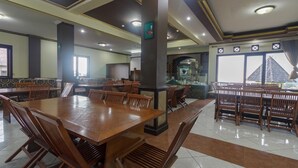 A spacious dining area with multiple wooden tables and chairs, a central pillar, and a view of the outside through large windows. - RedDoorz Syariah @ Cipanas (Cipanas)