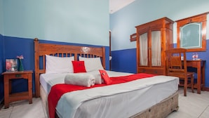A neatly made bed with white and red bedding, a wooden headboard, and a red pillow with a logo. There is a wooden nightstand with a lamp and a vase of flowers. A wooden wardrobe with a mirror and a chair is visible. - RedDoorz @ Genteng Surabaya 2 (Surabaya)