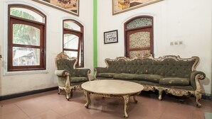 A room with a green upholstered sofa, a round coffee table, and two armchairs. There are large windows with wooden frames and a green vertical stripe on the wall. - RedDoorz @ Genteng Surabaya 2 (Surabaya)