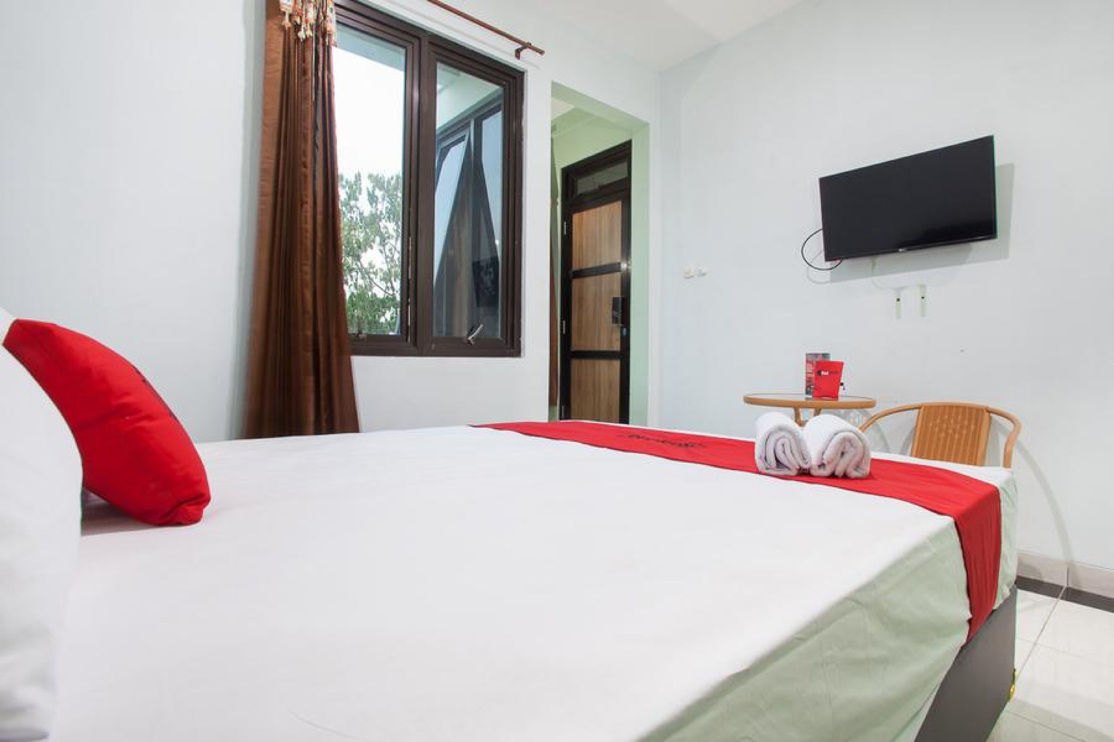 A hotel room with a bed, a TV mounted on the wall, a small table with a chair, and a window with curtains.