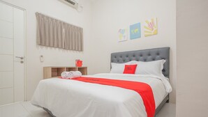 A neatly made bed with white and red bedding, a grey headboard, and a red pillow. There is a wooden nightstand with a red object on it, and a window with beige curtains. - RedDoorz near Ciputra Golf Surabaya (Surabaya)