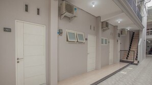 A hallway with three white doors, each marked with a number. There are air conditioning units mounted on the wall above the doors. The floor is tiled, and there is a staircase visible in the background. - RedDoorz near Ciputra Golf Surabaya (Surabaya)