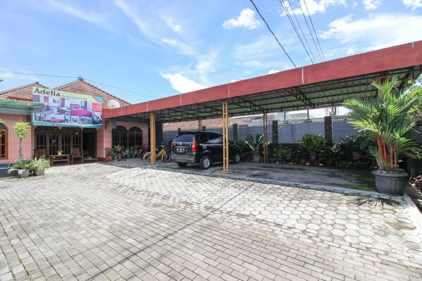 A covered outdoor area with a car parked under a shelter, potted plants, and a building with a sign advertising a homestay service. - RedDoorz near Candi Ratu Boko (Prambanan)
