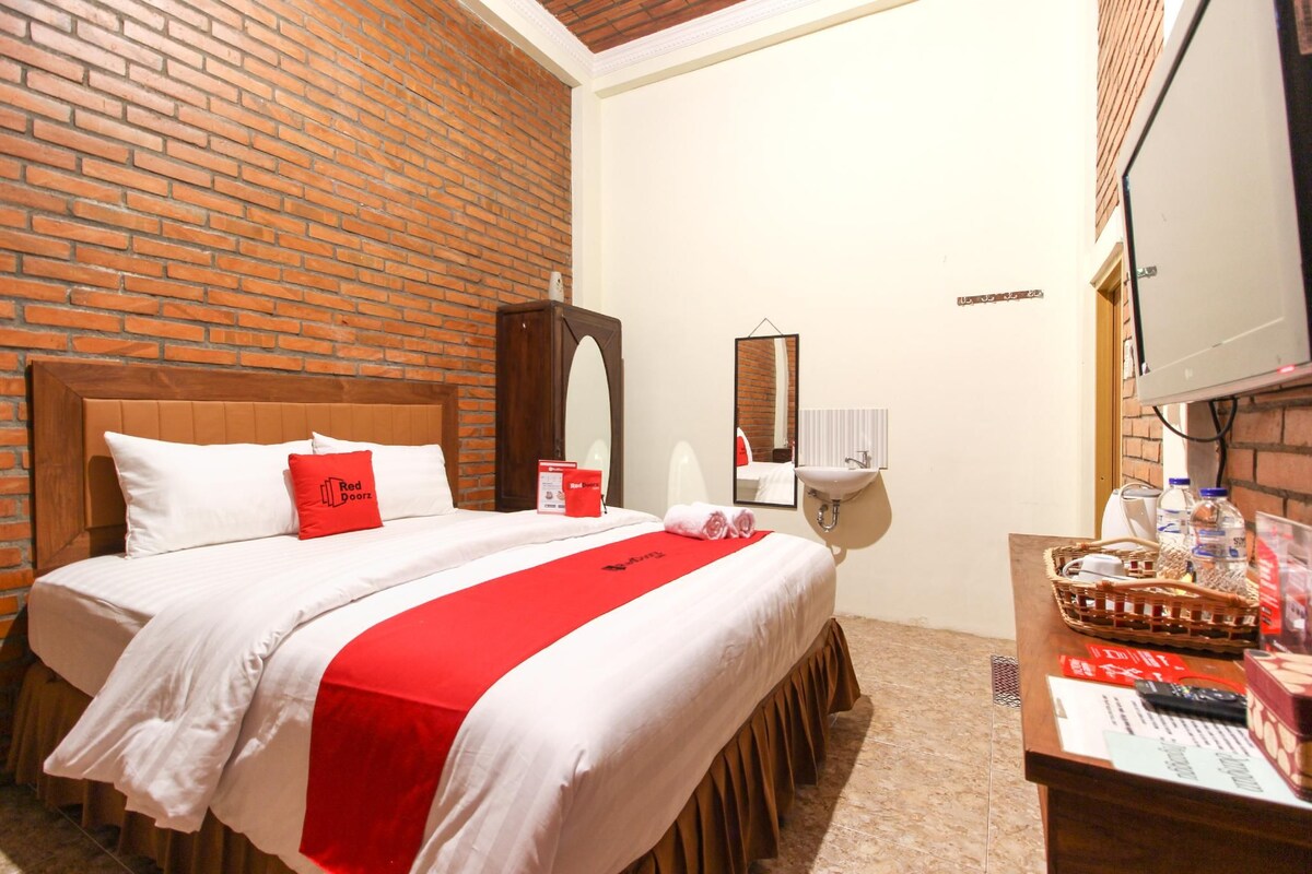 Habitació d'hotel amb paret de maó, llit de fusta amb roba de llit blanca i detalls vermells, un mirall, una tauleta de nit amb cistella i una televisió.