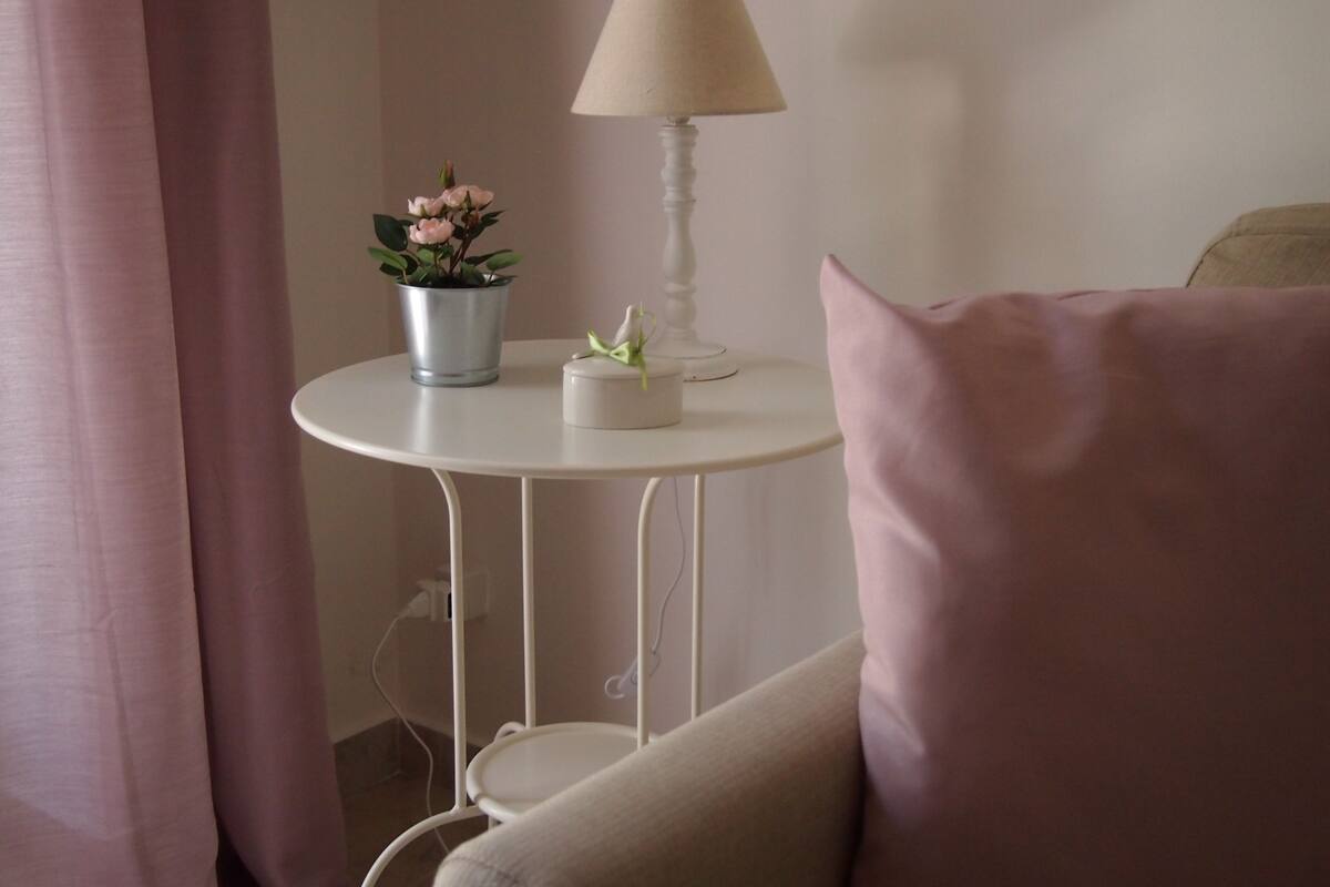 Una habitación con una mesa redonda blanca, una lámpara, una planta en maceta y un cojín rosa.