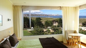 A hotel room with a large window offering a view of a coastal landscape, a bed with green bedding, a small table, and a chair. - Mantagua Village (Quintero)