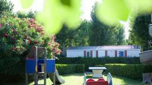 Un terrain de jeux avec un toboggan et une structure d’escalade, une table de pique-nique et un bâtiment aux volets rouges, le tout situé dans un cadre verdoyant.