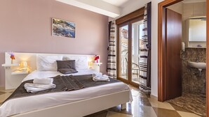 A neatly made bed with white linens and a brown throw, a nightstand with a lamp, a painting on the wall, and a bathroom visible through an open door. - Villa San Rocco Bed&Breakfast (Medulin)