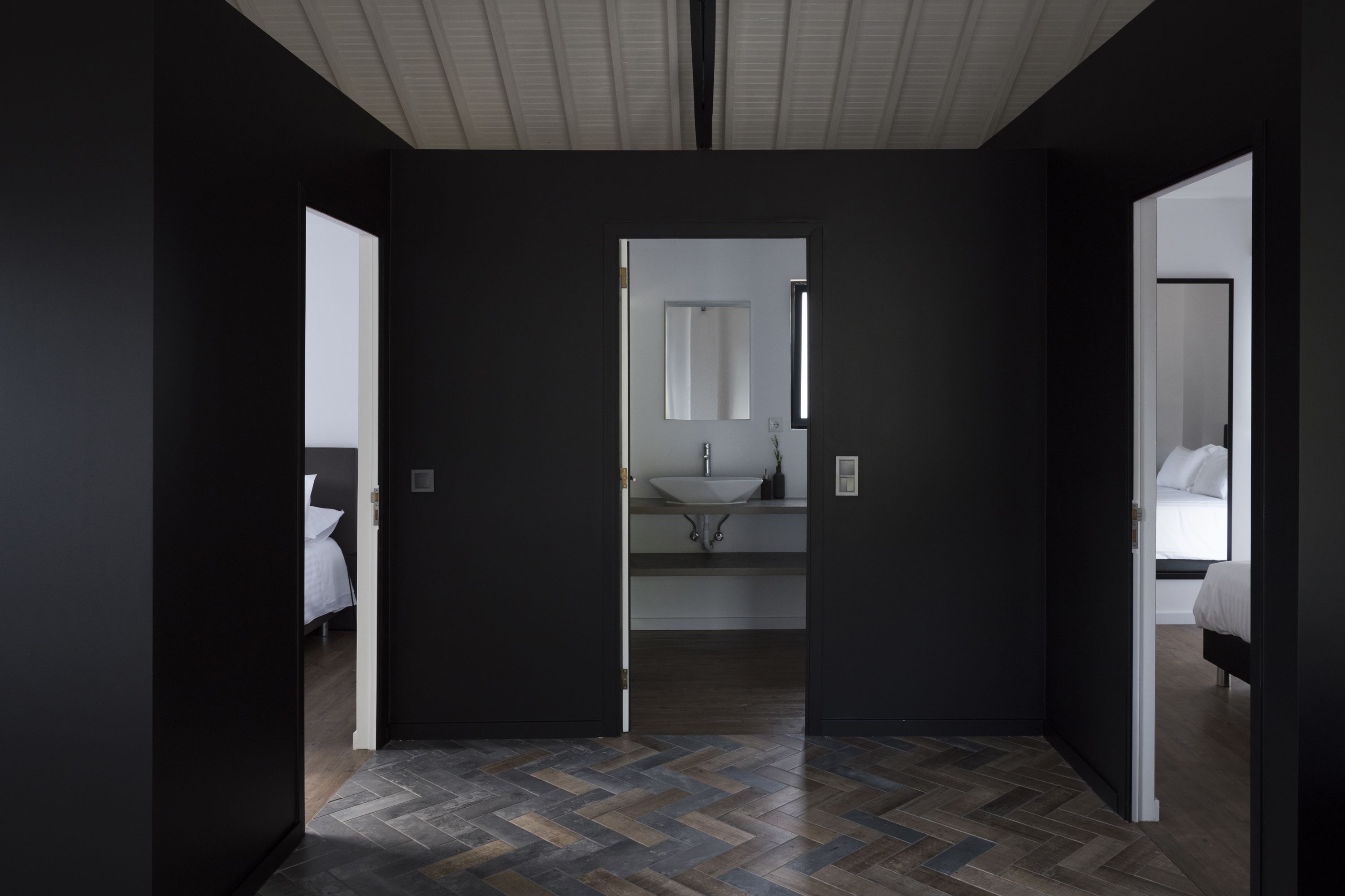 Una habitación de hotel moderna con interior oscuro, piso con estampado tipo espiga y un baño central con un espejo grande, además de una cama con ropa blanca.