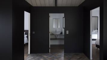 Una habitación de hotel moderna con interior oscuro, piso con estampado tipo espiga y un baño central con un espejo grande, además de una cama con ropa blanca.