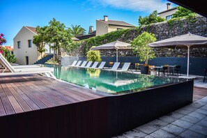 A modern outdoor pool area with lounge chairs, umbrellas, and a wooden deck. - Volcanic Charming House (Ribeira Grande)