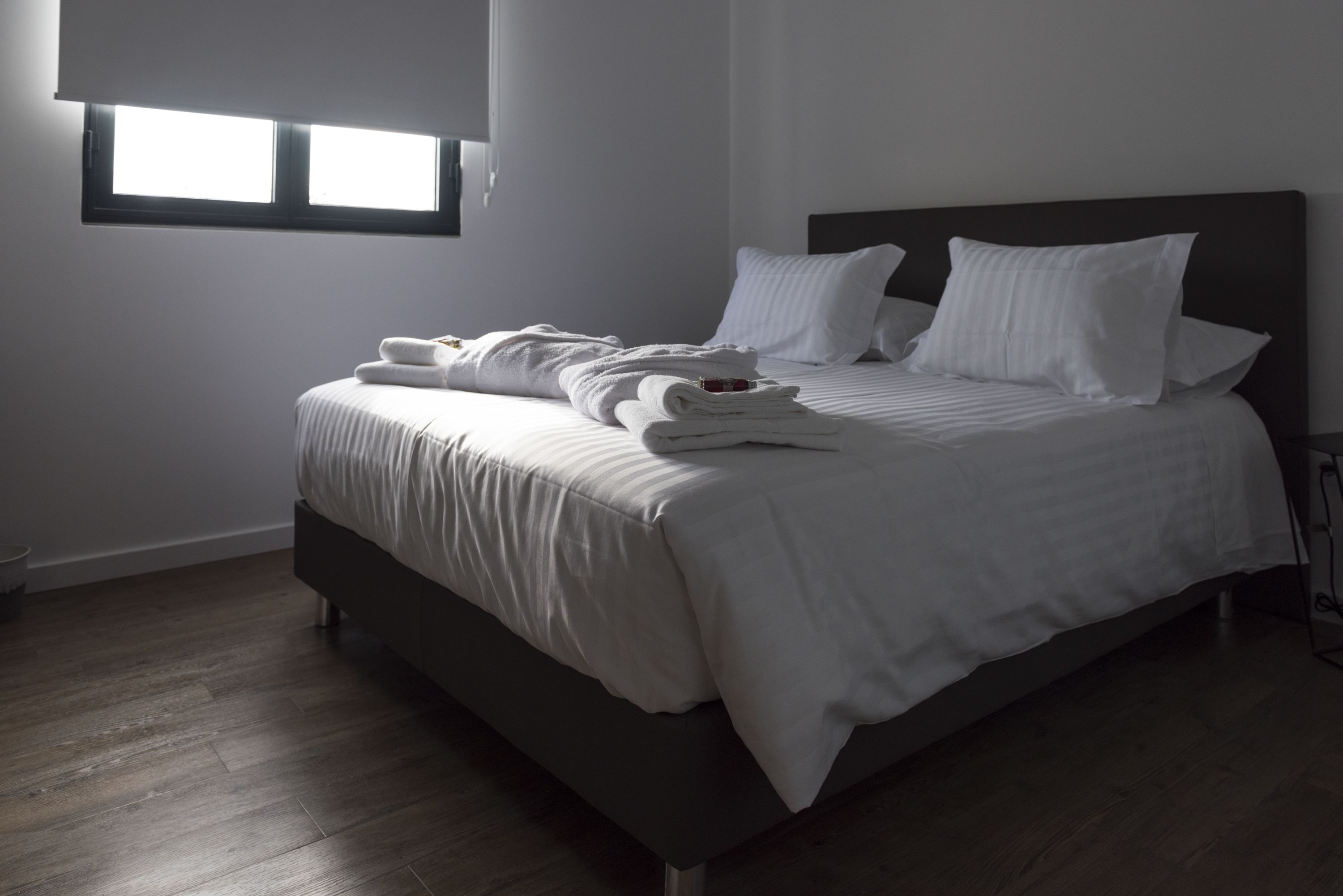 Una cama bien hecha con sábanas y almohadas blancas, un cabecero oscuro y una ventana con persianas.