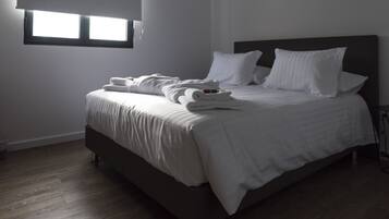A neatly made bed with white linens and pillows, a dark headboard, and a window with blinds.