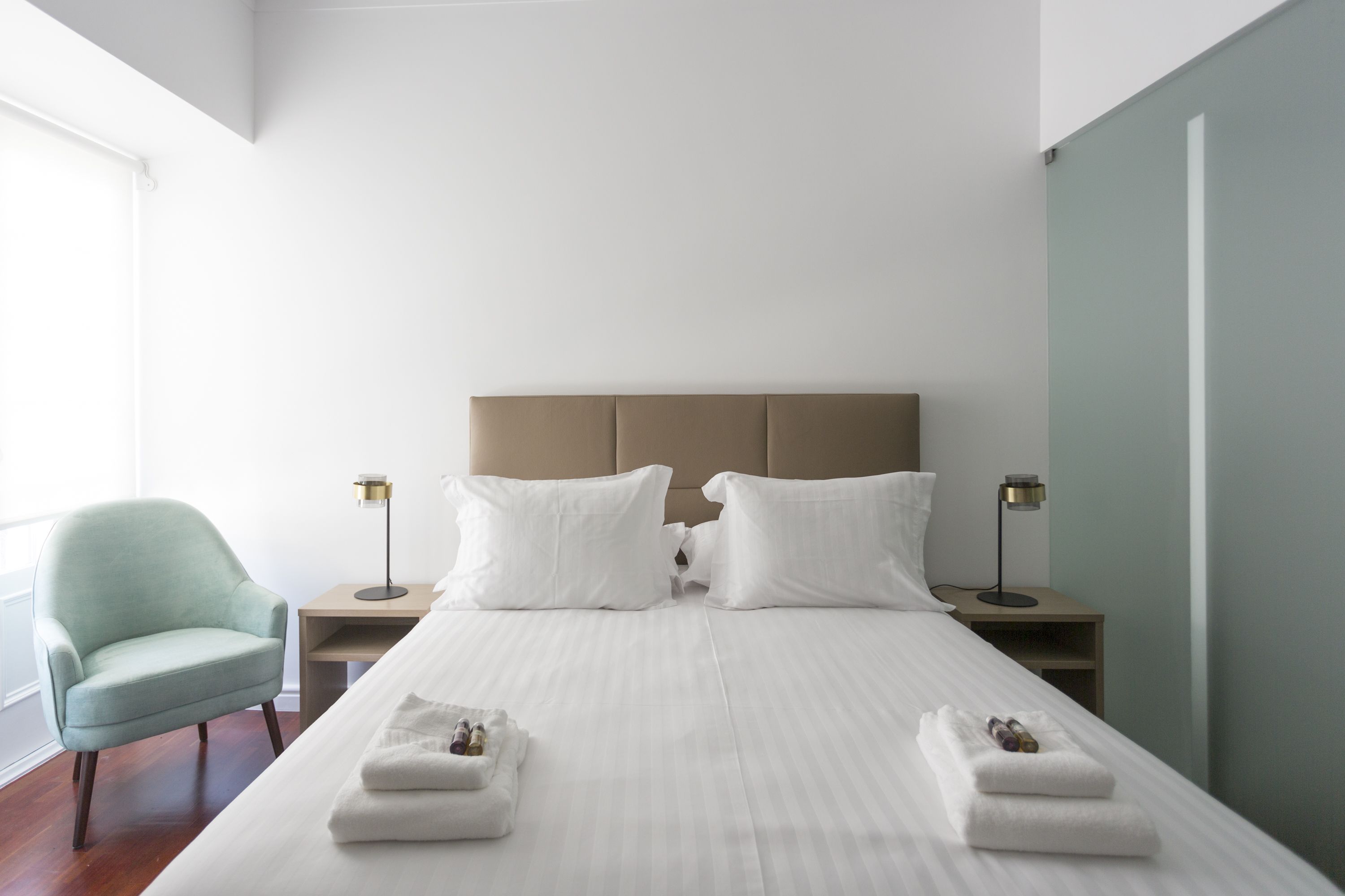 Een hotelkamer met een groot bed, twee nachtkastjes met lampen, een lichtgroene fauteuil en uitzicht buiten.