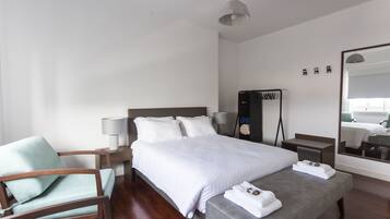 Ein modernes Hotelzimmer mit einem großen Bett, einer Sitzecke mit einem Sessel, einem Nachttisch mit Lampe, einem Spiegel und einem kleinen Tisch ebenfalls mit Lampe.