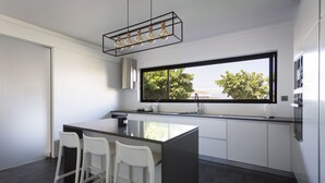 A modern kitchen with a large island, white bar stools, a window with a view, and a pendant light fixture. - Volcanic Charming House (Ribeira Grande)