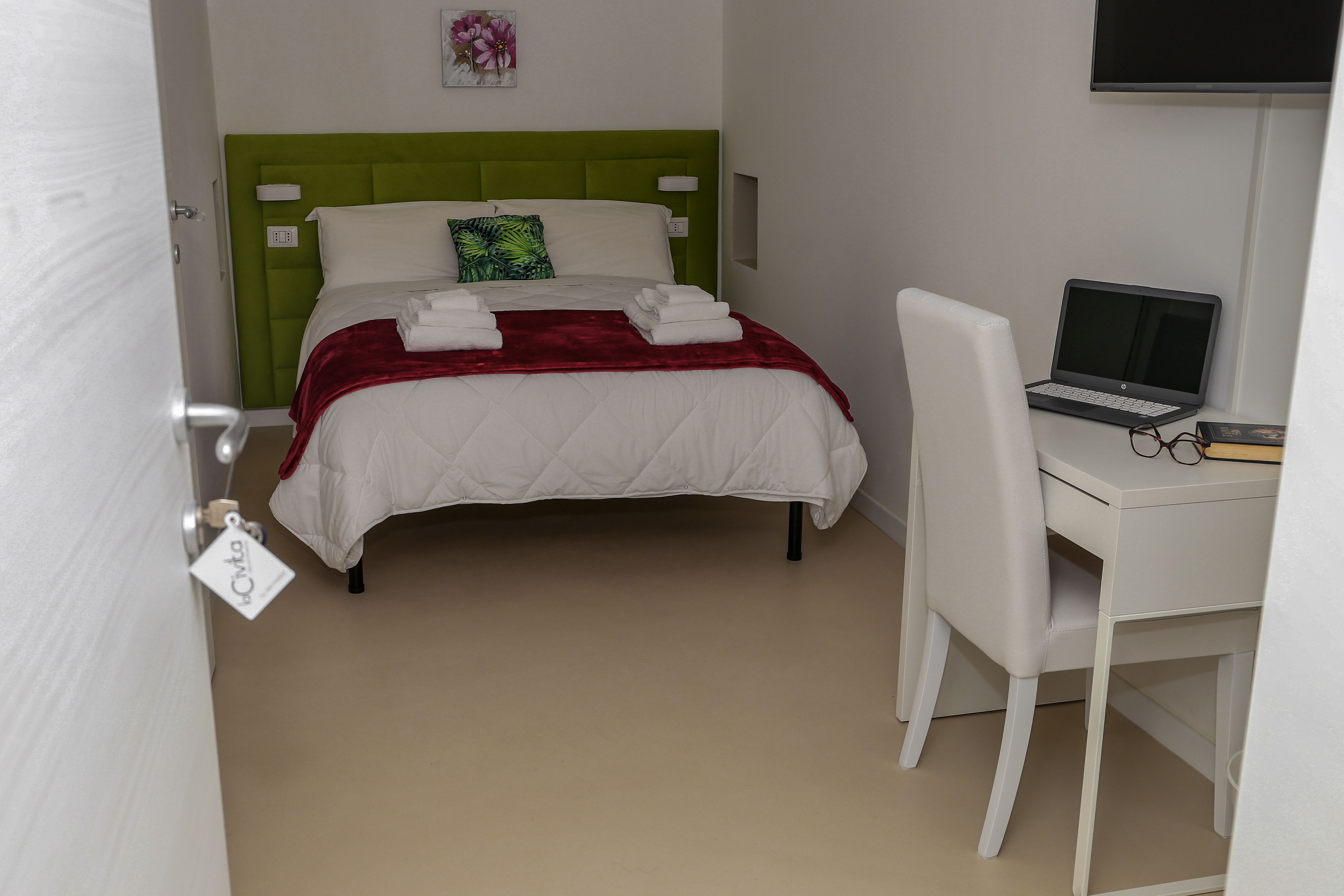 A neatly made bed with a green headboard, a white desk with a laptop, and a chair.