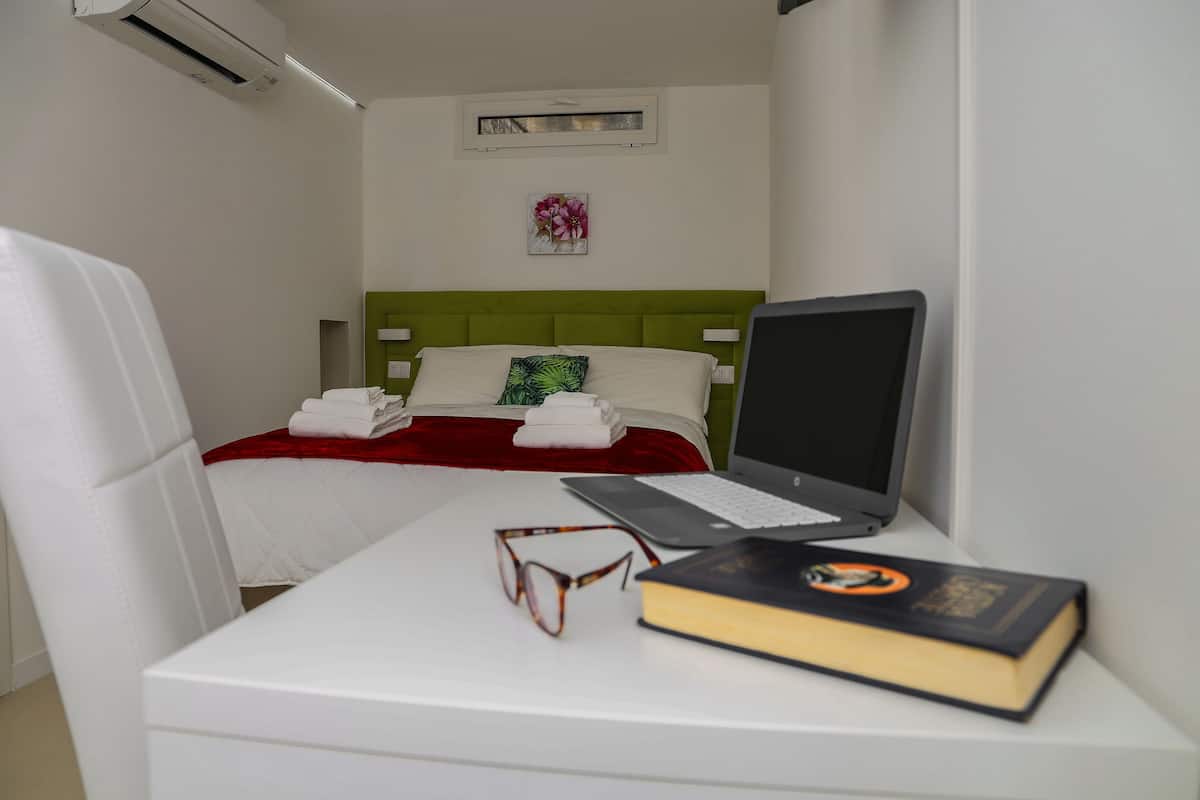 A hotel room with a bed, a desk with a laptop, a book, and a pair of glasses.