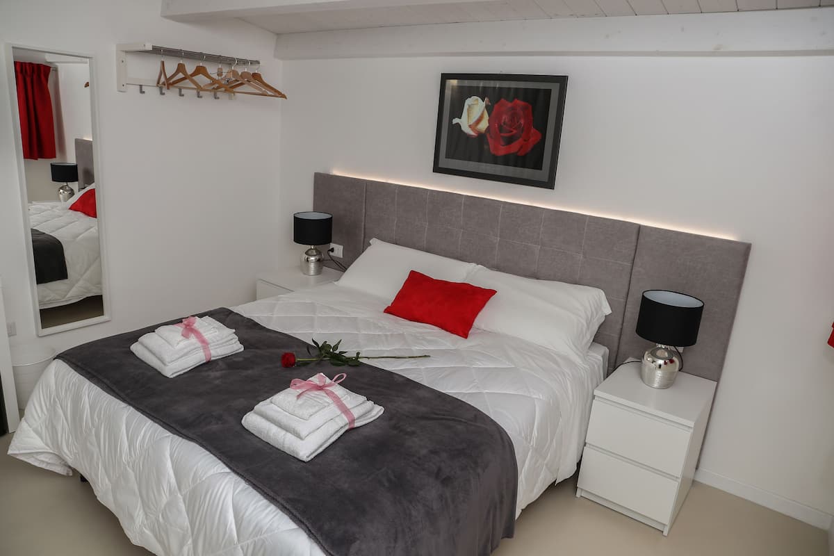 A neatly made bed with a grey blanket and red pillows, a bedside table with a lamp, and a framed picture of roses on the wall.