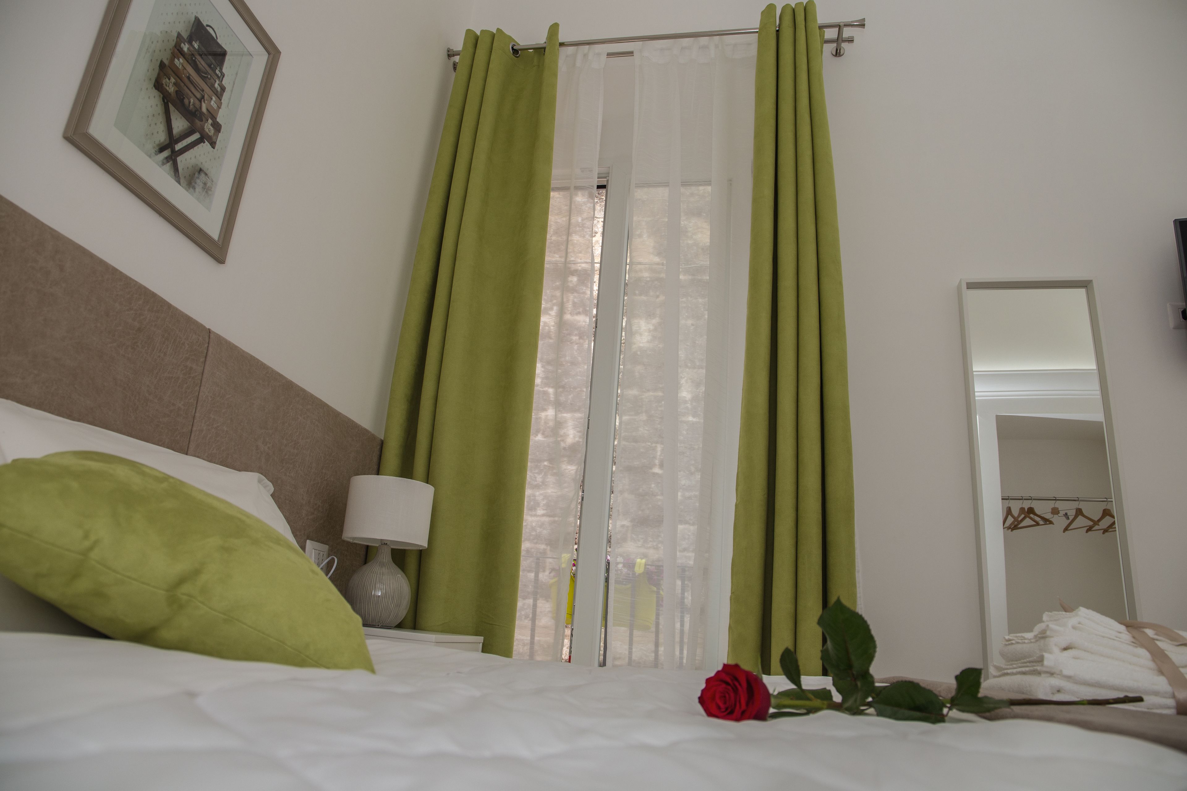 Una habitación de hotel con una cama, cortinas verdes, una lámpara blanca, un cuadro en la pared y un espejo con toallas.