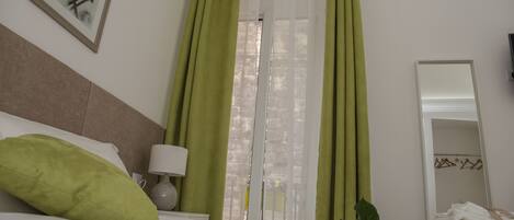 Una habitación de hotel con una cama, cortinas verdes, una lámpara blanca, un cuadro en la pared y un espejo con toallas.