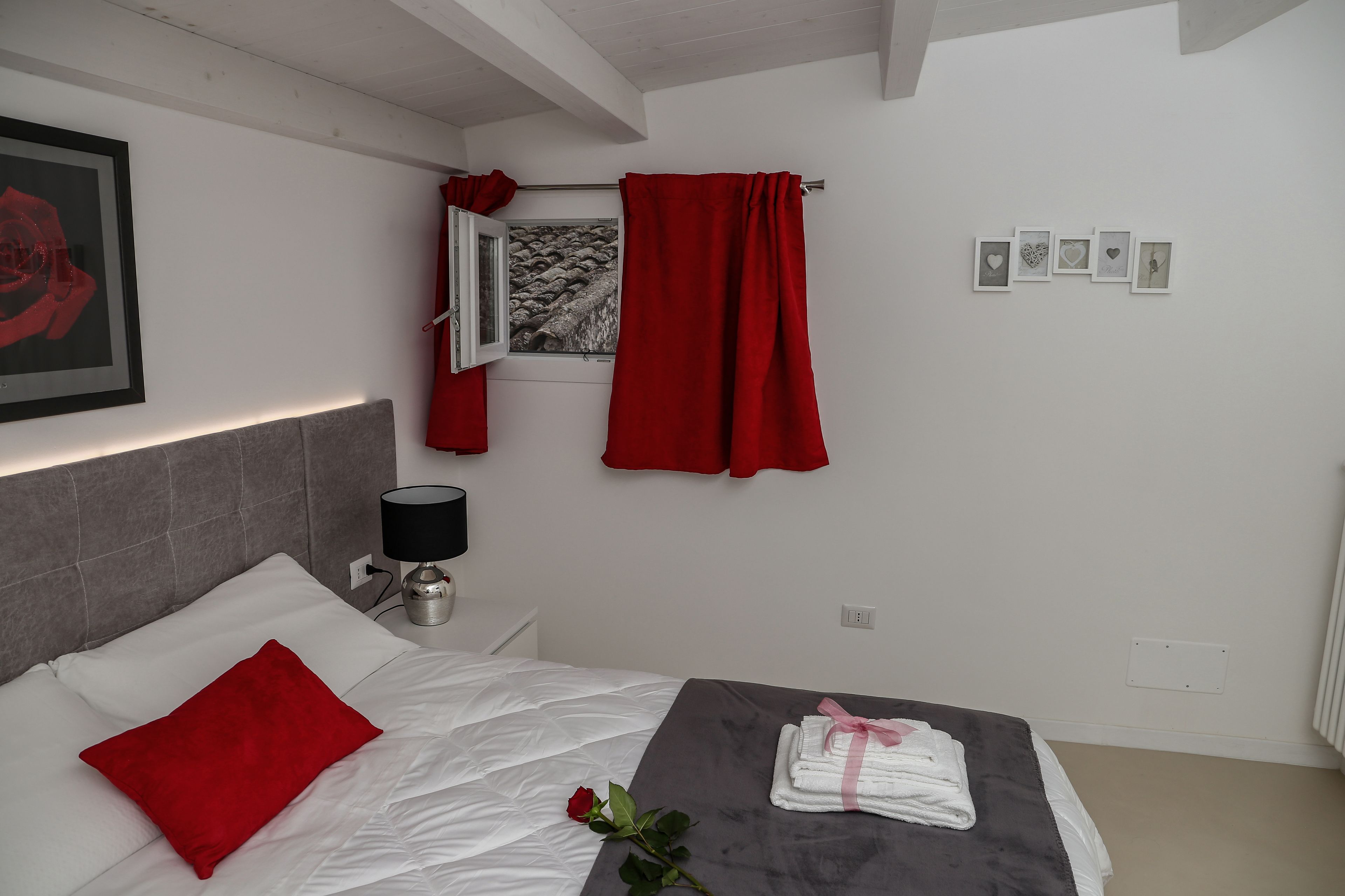 A hotel room with a bed, a nightstand with a lamp, a red rose on the bed, a towel on the floor, and a window with red curtains.