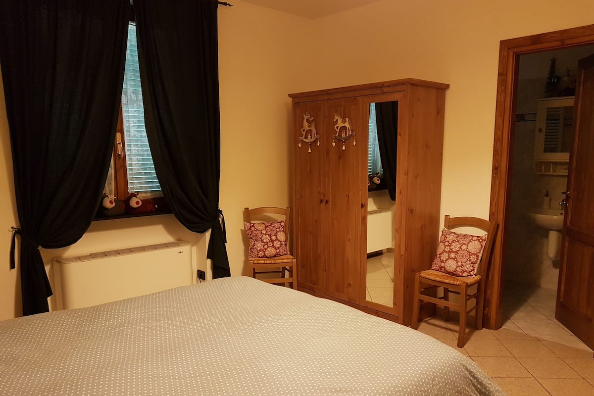 A bedroom with a bed, a wardrobe, two chairs, a window with curtains, and a bathroom visible through an open door.