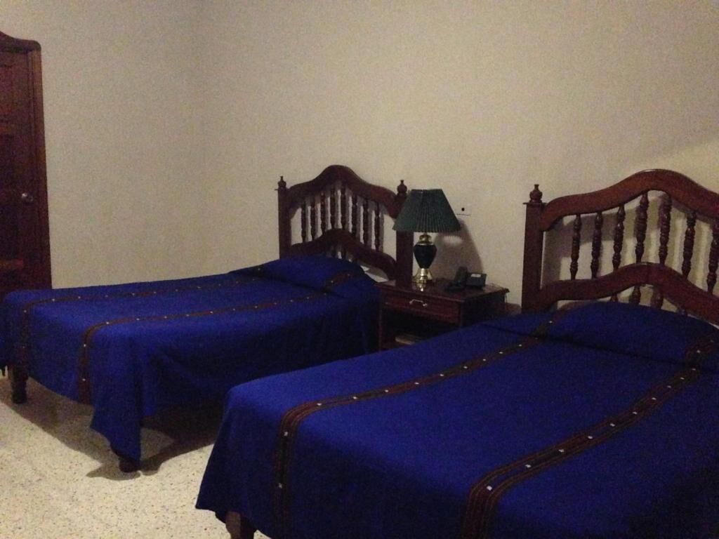 A room with two single beds, wooden headboards, a nightstand with a lamp, and a small table.