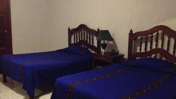 A room with two single beds, wooden headboards, a nightstand with a lamp, and a small table.
