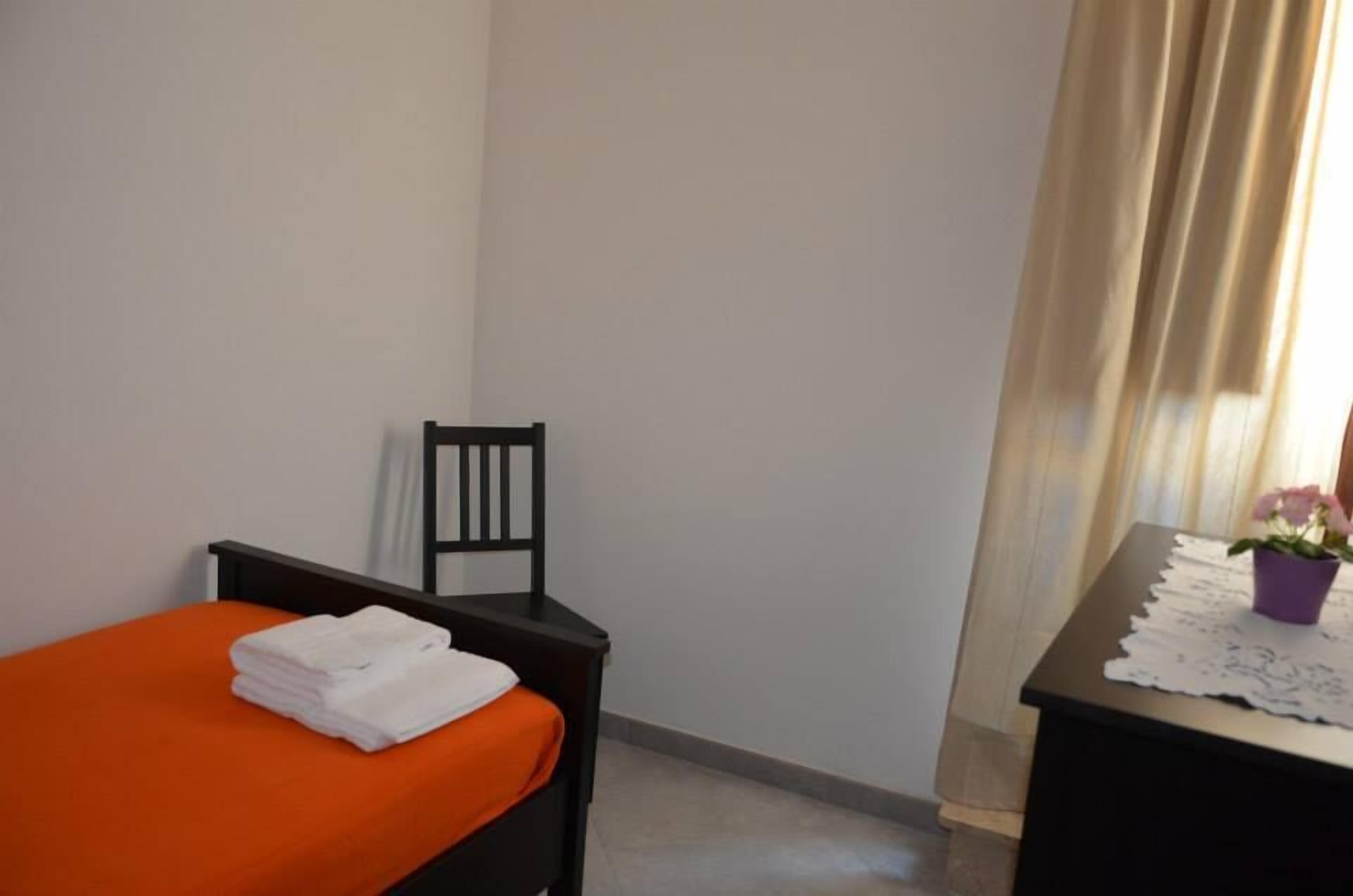 Une pièce simple, avec un lit, une chaise, une petite table recouverte d’une nappe en dentelle et une plante en pot.