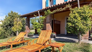 A patio with wooden lounge chairs and a small table, surrounded by a garden with trees and a building with a tiled roof.