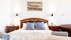 A neatly made bed with a patterned bedspread, two pillows, and a headboard. There are two bedside tables with lamps, a brown leather chair, and a wall-mounted painting.