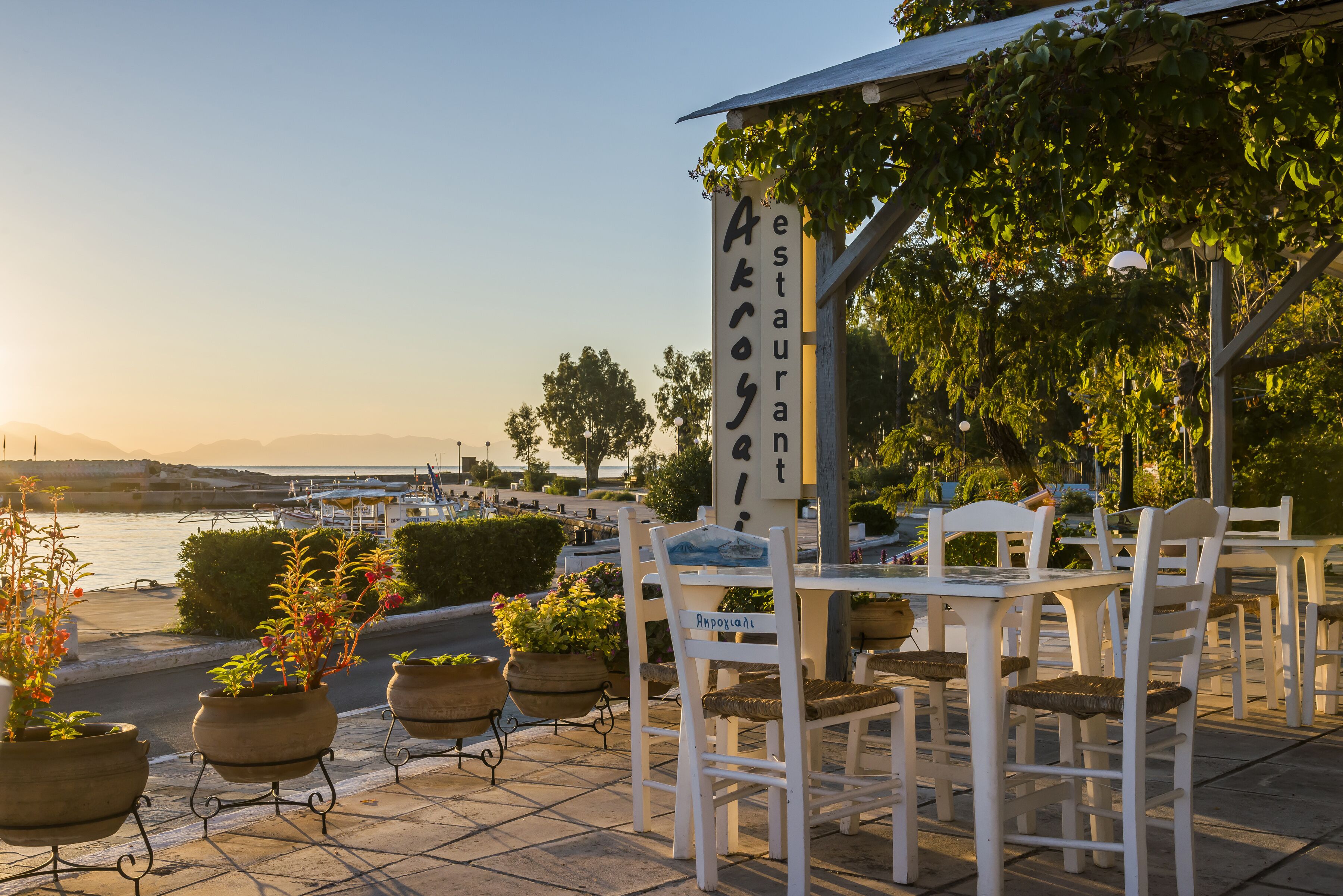 An outdoor seating area with white tables and chairs, potted plants, and a sign for "Akroos Restaurant" overlooking a body of water.