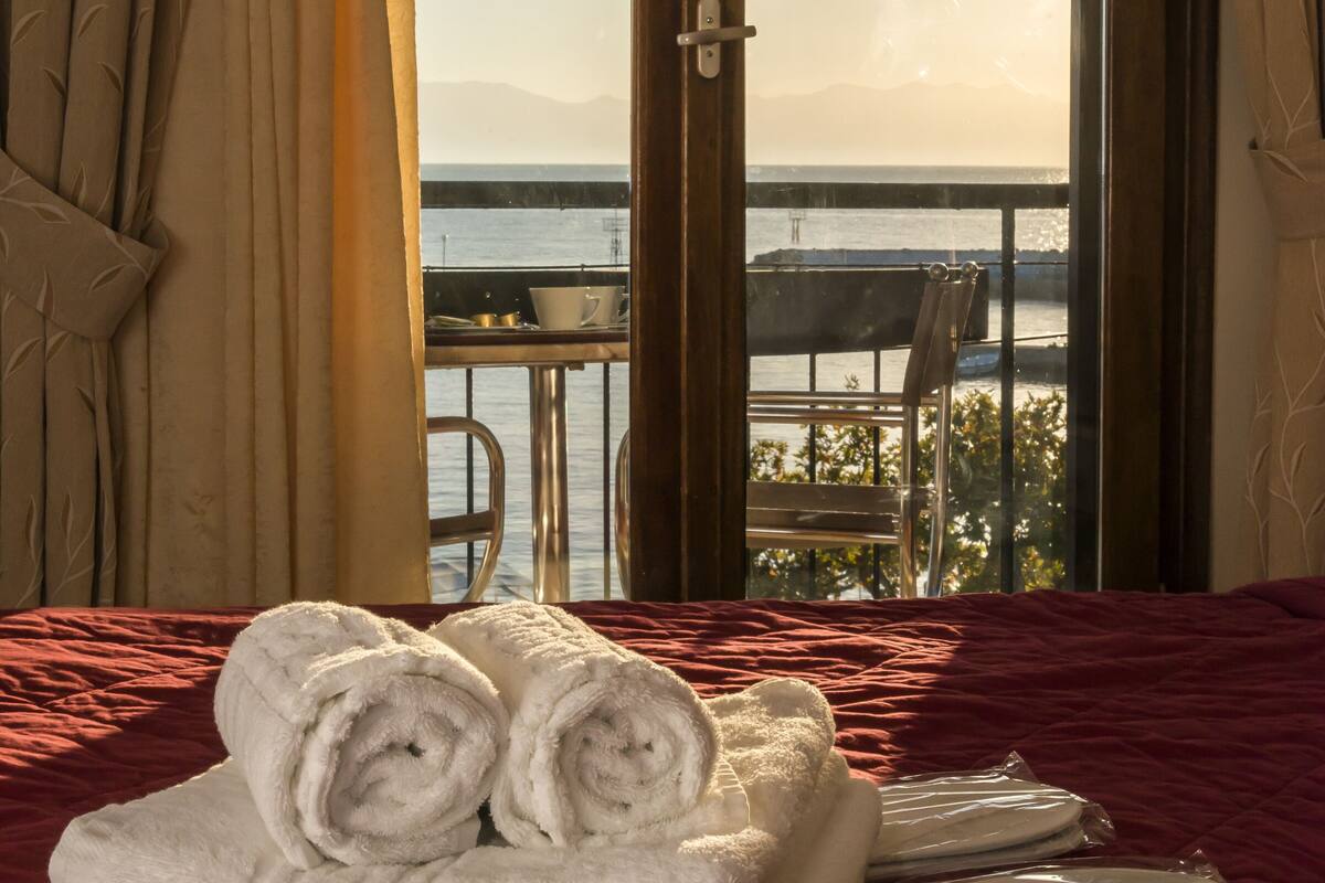 A hotel room with a bed, a set of towels, a dining table with plates, and a view of the sea through a balcony door.